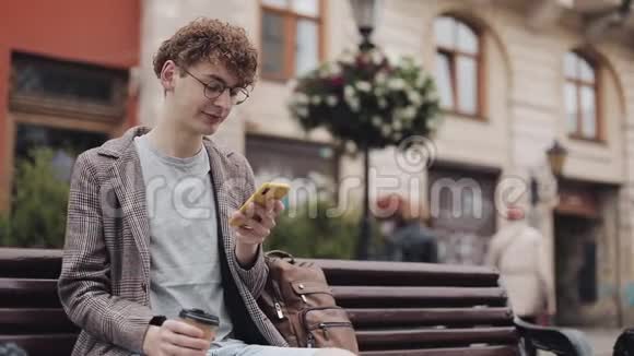 戴眼镜夹克和包躺着的年轻嬉皮士用智能手机侧视在屏幕上滚动视频的预览图