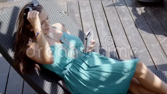 穿着蓝色衣服的女人在躺椅上休息用智能手机视频的预览图