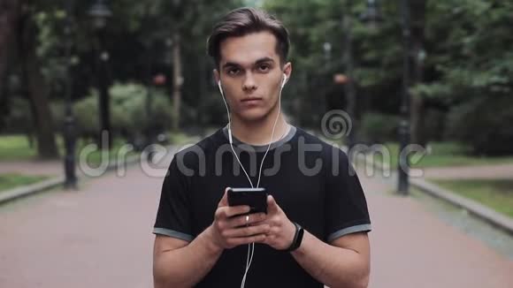 有魅力的年轻人在耳机和智能手机中站在公园概念健康生活方式的肖像视频的预览图