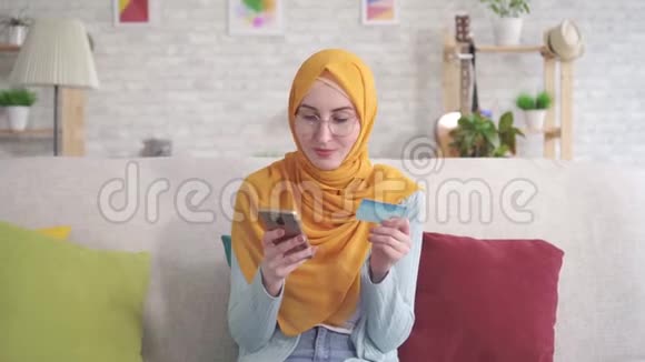 肖像积极的年轻穆斯林妇女戴着头巾微笑着拿着智能手机和银行卡坐在客厅的沙发上视频的预览图