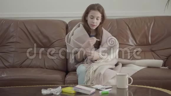 肖像小病女坐在沙发上药片药丸和杯子放在桌子上女性看温度计概念视频的预览图