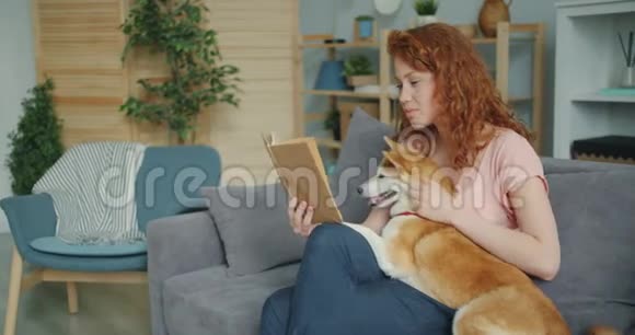 微笑的女孩读着有趣的故事抚摸着坐在家里沙发上的小狗视频的预览图