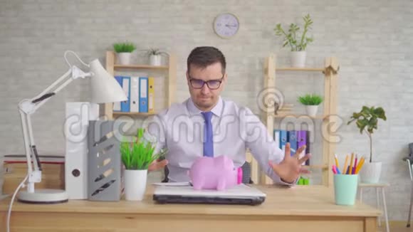 穿着衬衫的男人坐在办公室的笔记本电脑旁手里拿着一个储蓄罐视频的预览图