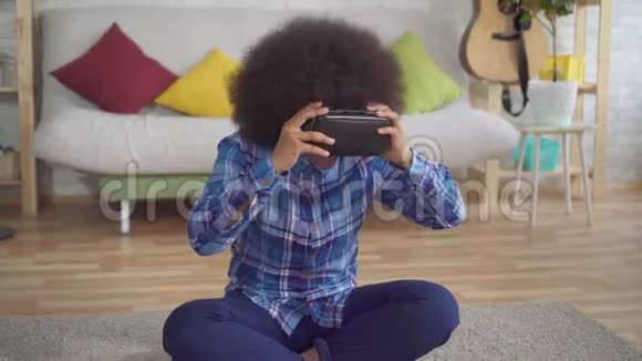 表现积极的非裔美国年轻女性在客厅使用VR眼镜和虚拟技术视频的预览图