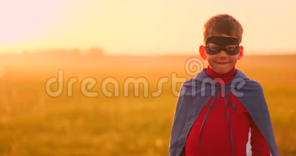 一个男孩戴着超级英雄的面具看着日落时站在田野里的摄像机的肖像视频的预览图