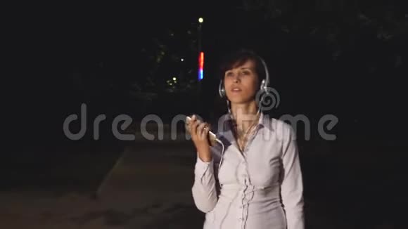 戴着耳机和智能手机的年轻女孩在一个城市公园里听音乐跳舞和微笑开朗活泼视频的预览图