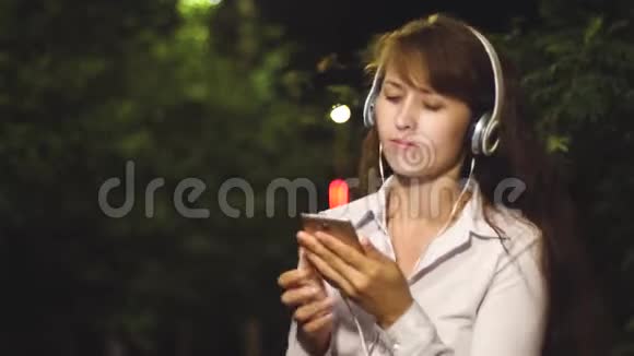 开朗的女孩在夜城旅行戴着耳机和智能手机的年轻女孩正在听音乐跳舞和视频的预览图