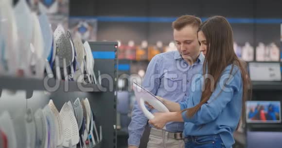 年轻的已婚夫妇选择在电器商店的一个新家买熨斗一个女人握着她的手看着视频的预览图