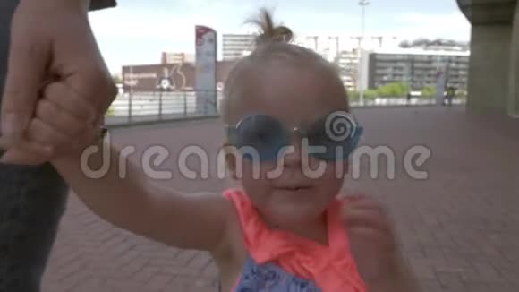 一个穿着非常时髦的蓝色圆形太阳镜的漂亮头发的女婴牵着母亲的手走着视频的预览图