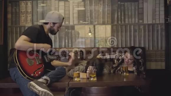 年轻有胡子的男人在酒吧弹吉他他的朋友坐在附近在酒吧休闲男人和女孩视频的预览图
