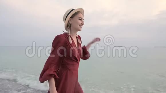 日落时分一个年轻迷人的微笑女孩穿着红色的长裙戴着草帽在风和日丽的天气里沿着海滨散步视频的预览图