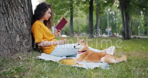 放松的女孩在公园里和狗坐在草坪上喝咖啡和看书视频的预览图