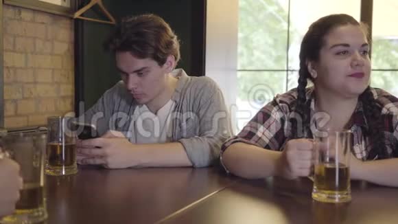 丰满的女孩坐在酒吧的桌子上和朋友喝啤酒年轻人一边打手机一边发短信视频的预览图