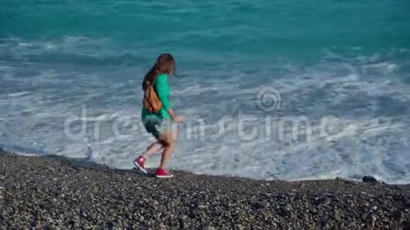 快乐的年轻女子穿着绿色夹克牛仔短裤和运动鞋背着背包在温暖的阳光下沿着大海散步视频的预览图