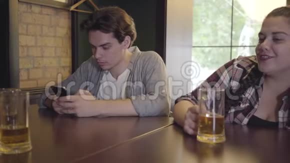 胖女孩和朋友一起喝啤酒坐在酒吧的桌子旁年轻人一边打手机一边发短信视频的预览图
