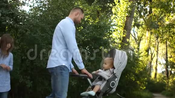 年轻幸福的一家人在夏季公园散步爸爸带着婴儿车妈妈带着女儿走视频的预览图