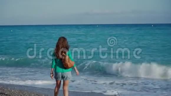 快乐的年轻女子穿着绿色夹克牛仔短裤和运动鞋背着背包在温暖的阳光下沿着大海散步视频的预览图