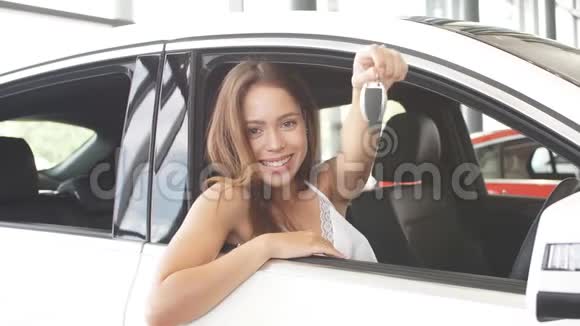 快乐的白种人年轻女司机坐在白色的车上在汽车陈列室里展示汽车钥匙视频的预览图