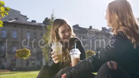 快乐的白种人大学女生在休息的时候坐在草坪上喝着牛奶鸡尾酒和说话视频的预览图