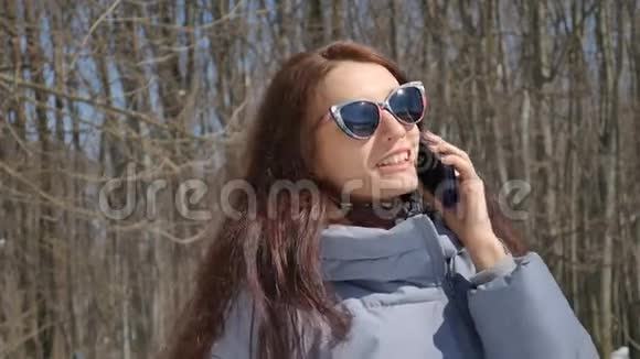 漂亮的黑发女孩穿着时髦的太阳镜在公园里的树上打电话给一个在户外使用蓝色智能手机的人视频的预览图