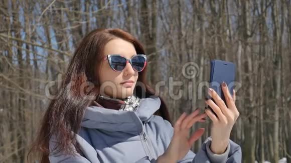 漂亮的黑发女孩穿着时髦的太阳镜在公园的户外用蓝色智能手机拍照视频的预览图