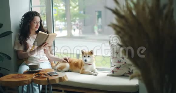 漂亮的女孩抚摸着窗台咖啡馆里的石巴狗和看书视频的预览图
