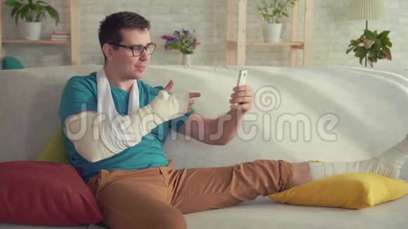 一个胳膊和腿断了的年轻人坐在沙发上视频通话视频的预览图