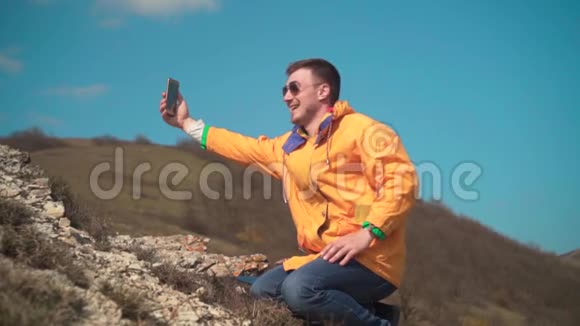 一个穿着黄色夹克蓝色牛仔裤和眼镜的年轻人坐在山上欣赏风景在视频上交谈视频的预览图