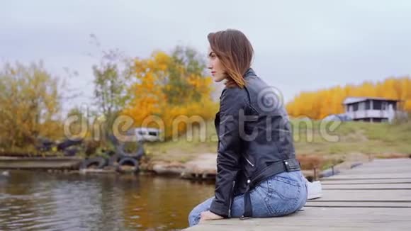 一个穿着皮夹克的女孩坐在木桥边晃着腿那个女孩坐在码头上河流视频的预览图