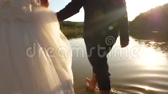 新娘和新郎赤脚在河岸上的水上行走特写镜头一对相爱的夫妇手牵手走在海滩上视频的预览图