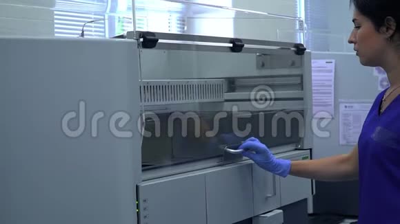 穿着蓝色制服和橡胶手套的年轻女子打开了实验室工作机器的门看着里面视频的预览图