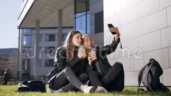 两个带着新鲜果汁的白种人少女坐在草坪上用手机自拍视频在户外聊天视频的预览图