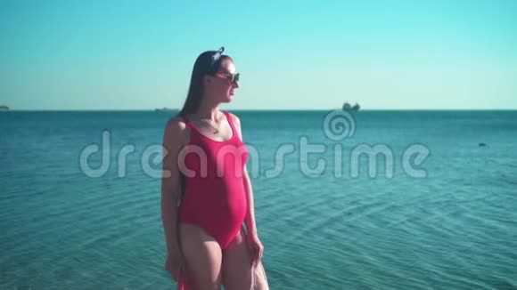 一个穿着红色一件泳衣和眼镜的怀孕女孩站在海边对着镜头微笑视频的预览图