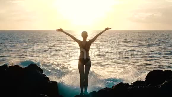 日落时分一位妇女举起手臂在空中观察巨大的海浪撞击岩石和喷洒视频的预览图