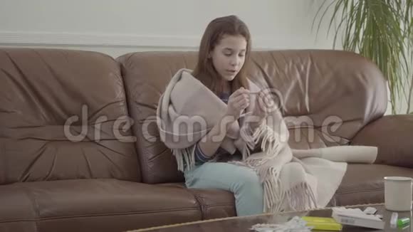 肖像小生病的女孩坐在沙发上药片药丸和杯子放在桌子上女性看温度计概念视频的预览图