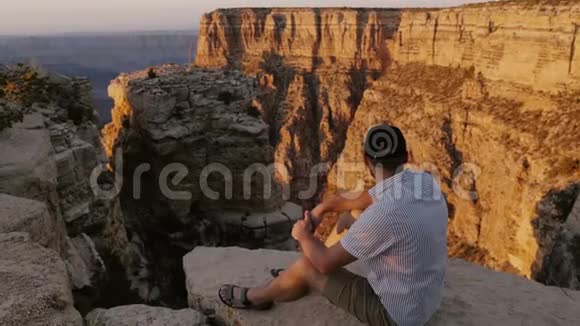 大峡谷上坐在大岩石上观看日落景色的年轻游客正在观看高角度摄像机视频的预览图