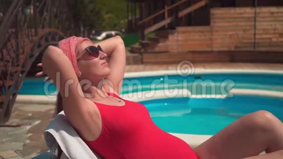 一个怀孕的女孩穿着红色的一件泳衣和眼镜双手放在她的头下靠近游泳池视频的预览图
