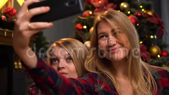 两个漂亮的年轻女孩或女人坐在新年或圣诞树附近用手机自拍视频的预览图