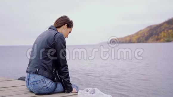 一个穿着皮夹克的女孩坐在木桥边晃着腿那个女孩坐在码头上河流视频的预览图