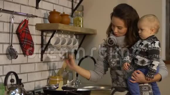 漂亮的年轻女人怀里抱着一个孩子在现代厨房准备早餐女士把盐倒进煎锅里视频的预览图