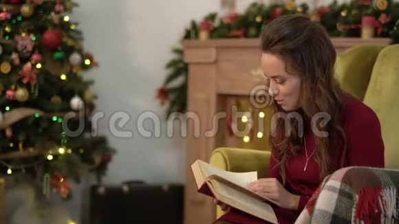 可爱迷人的年轻女人坐在圣诞树旁边的椅子上看书视频的预览图