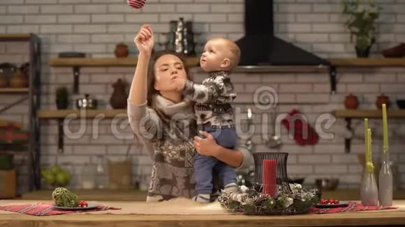 漂亮的年轻女子把婴儿抱起来站在现代厨房里向儿子展示鲜红的圣诞树玩具视频的预览图