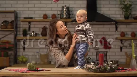 漂亮的年轻女子把婴儿抱起来站在现代厨房里向儿子展示鲜红的圣诞树玩具视频的预览图
