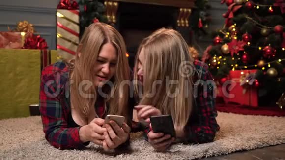 两个穿着格子红衬衫的漂亮金发妹妹躺在新年或圣诞树附近互相展示视频的预览图