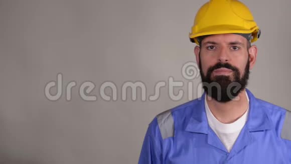 戴着头盔和蓝色制服的胡子工人在灰色背景下看着摄像机视频的预览图