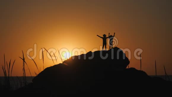 孩子们在夕阳下剪影戴帽檐的男孩站在岩石上石头躺在海边视频的预览图