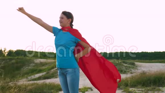 美丽的女孩扮演超级英雄穿着红色斗篷站在田野上斗篷随风飘扬年轻女孩穿着红色的衣服走视频的预览图