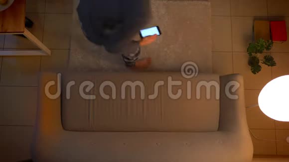 年轻男性坐在沙发上用手机上的一个应用程序作为遥控器打开电视的特写镜头视频的预览图