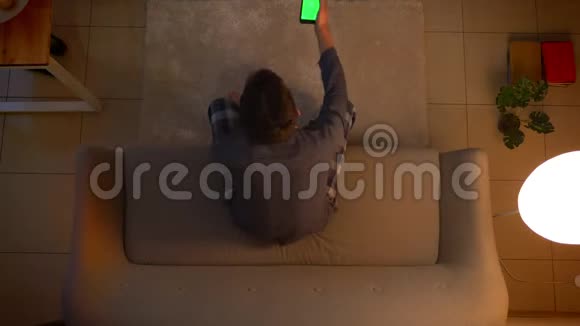 特写镜头拍摄年轻男性看电视和使用手机应用程序与绿色屏幕作为遥控器坐着视频的预览图