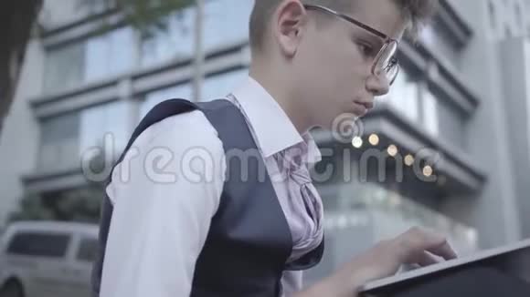 英俊的穿着漂亮的男孩戴着眼镜坐在街上用笔记本电脑做特写严肃的孩子像成年人一样视频的预览图